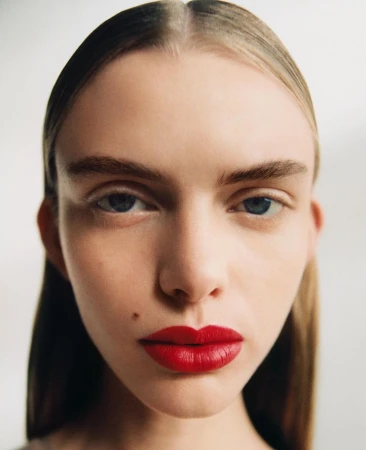Close-up portrait of a woman with slicked-back blonde hair, blue eyes, and bold red lipstick against a plain white background.