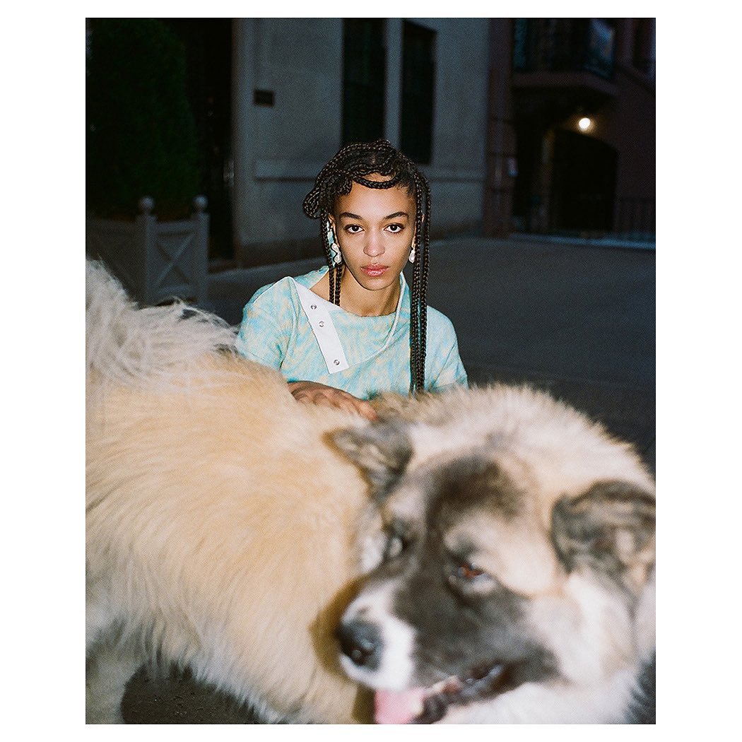 Adult female poses with large dog on city street, wearing braided hair and tie-dye top, urban setting with buildings in background.