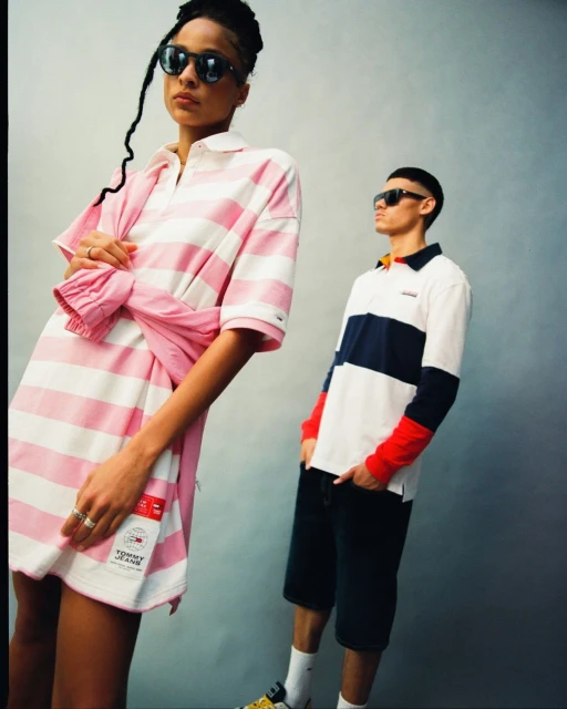 Two fashion models pose in an indoor setting. The woman wears a pink and white striped rugby dress with sunglasses, while the man wears a striped rugby shirt.