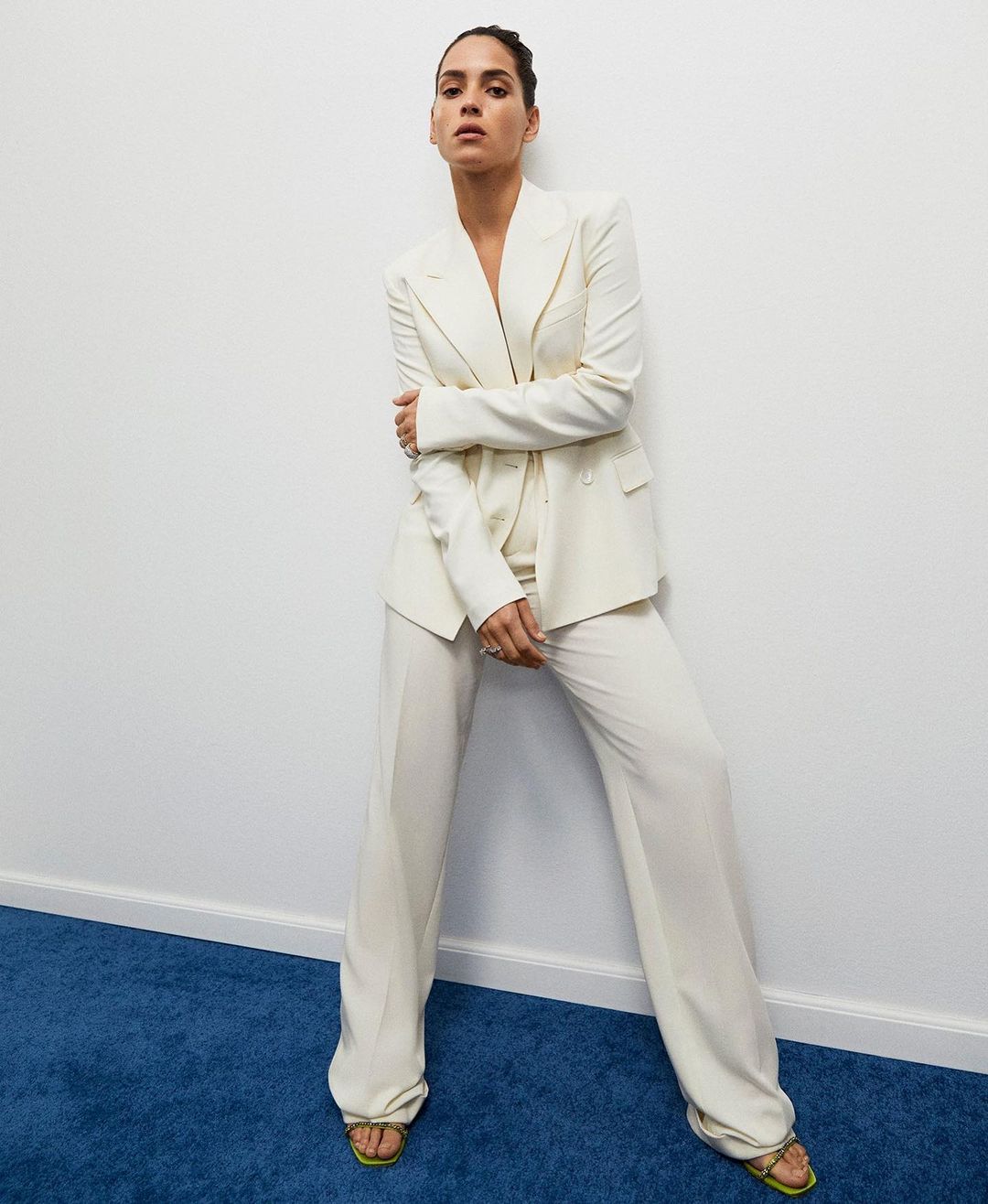 An adult woman poses indoors wearing a cream-colored pantsuit and matching sandals against a white wall and a blue carpeted floor.