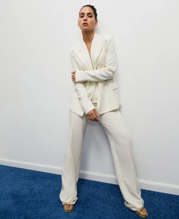 An adult woman poses indoors wearing a cream-colored pantsuit and matching sandals against a white wall and a blue carpeted floor.