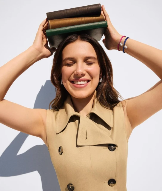 Smiling woman, Millie Bobby Brown, in a tan sleeveless trench coat, balancing three books on her head against a white background.