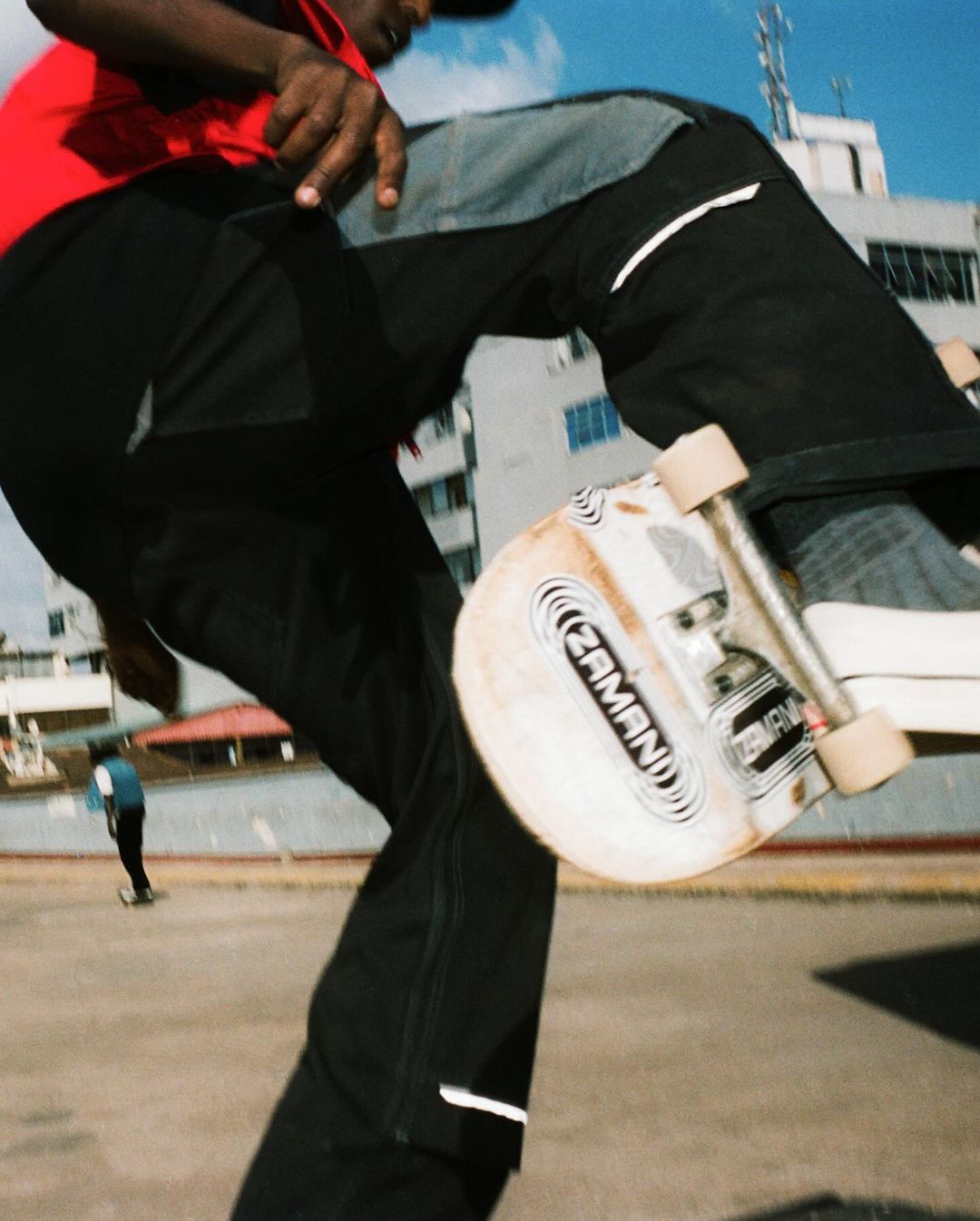 A person rides a skateboard on a concrete surface near a building, captured from a low angle with a clear blue sky backdrop.