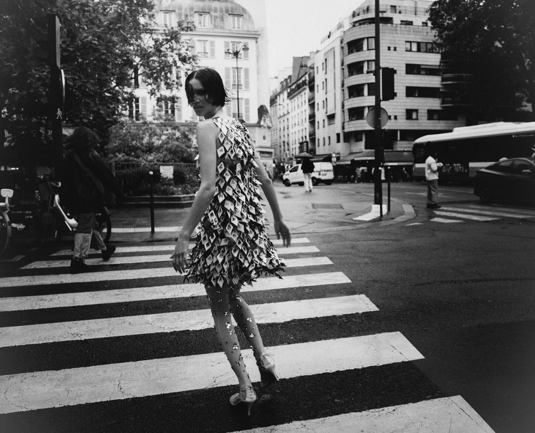 Monochrome shot of a woman in a diamond-pattern dress walking on a zebra crossing in a city street with buildings and traffic.