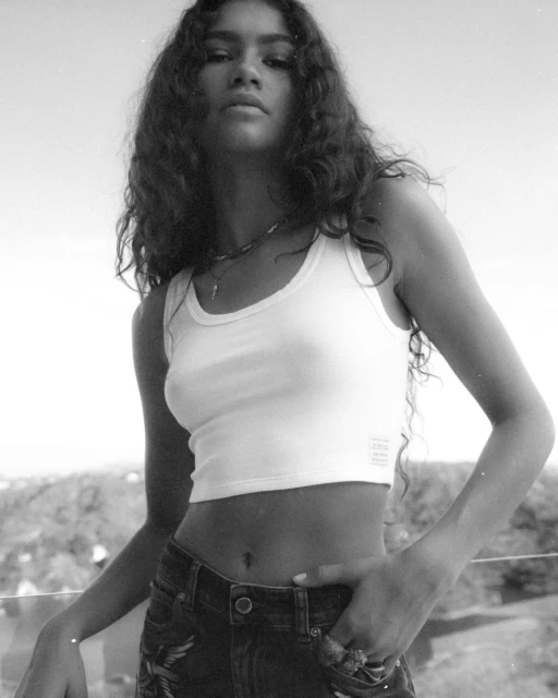Black and white portrait of Zendaya in a white tank top and dark jeans, striking a pose in front of an outdoor cityscape backdrop.