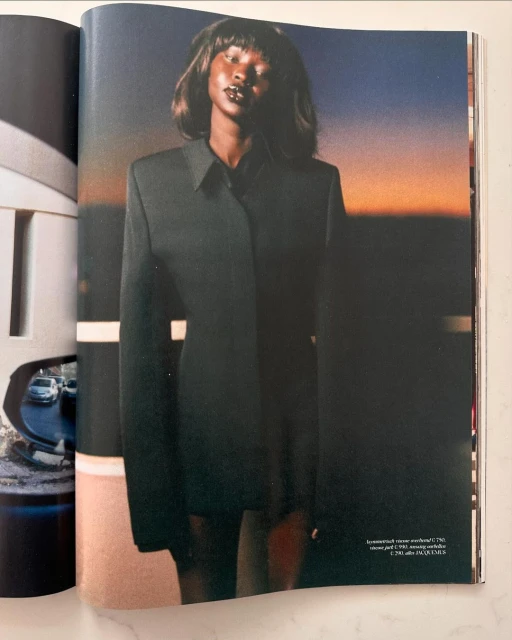 An African American woman stands in a black blazer against a sunset background in a fashion editorial shot for print media.