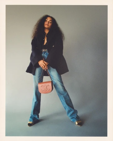 Woman with long dark hair wearing a dark jacket and blue jeans, holding a tan handbag against a plain grey backdrop.