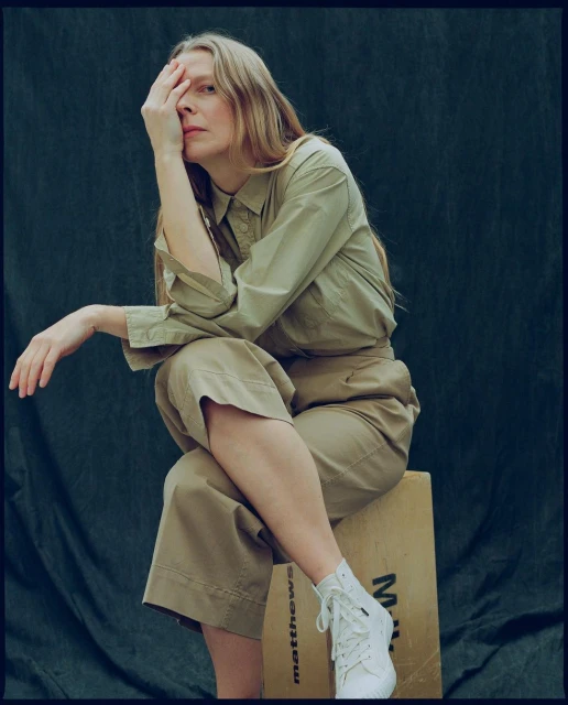 Woman in stylish casual outfit posing in photo studio, seated on wooden box with fair skin and blonde hair, eye partially covered.