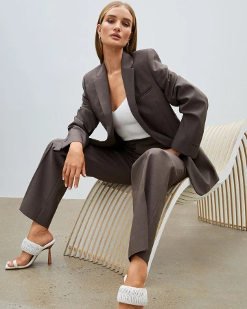 A stylish adult woman in a brown suit and white heels poses in a studio setting on a modern chair, looking confidently at the camera.