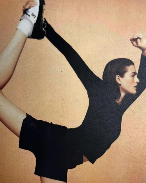 Athletic woman in black workout attire balances on one leg while holding the other up, posing against a tan-colored backdrop.