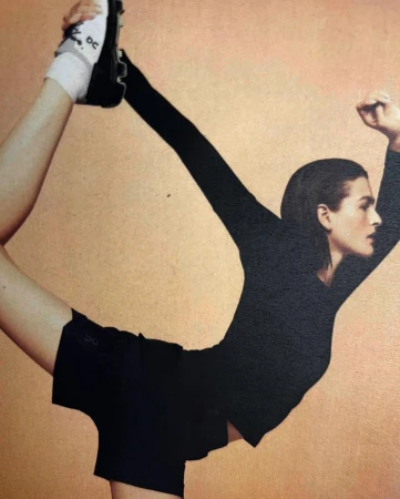 Athletic woman in black workout attire balances on one leg while holding the other up, posing against a tan-colored backdrop.