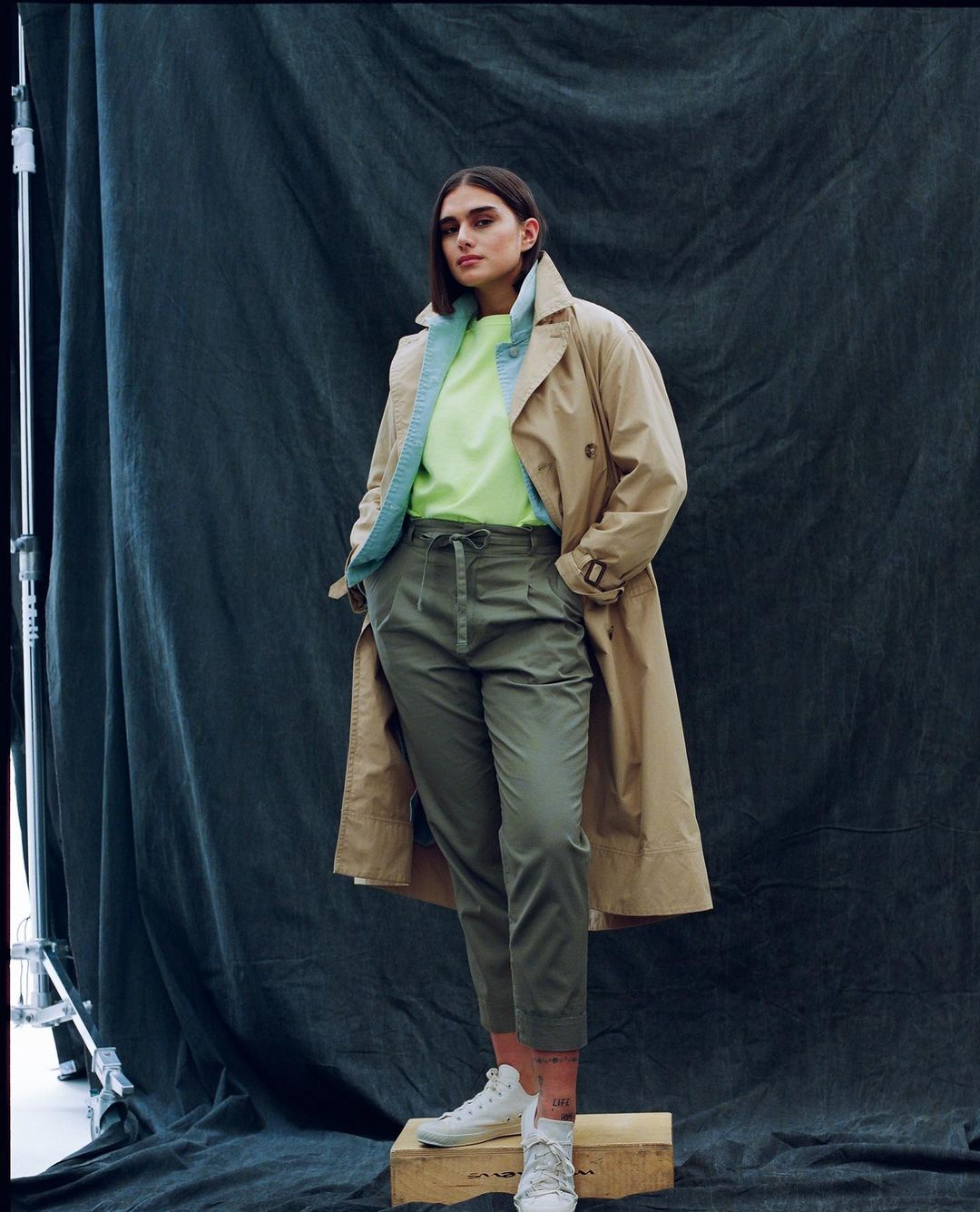 Fashionable young adult woman in a tan trench coat, lime-green top and olive green trousers poses in front of a dark backdrop.