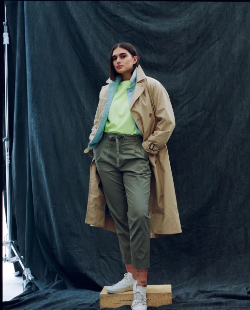 Fashionable young adult woman in a tan trench coat, lime-green top and olive green trousers poses in front of a dark backdrop.