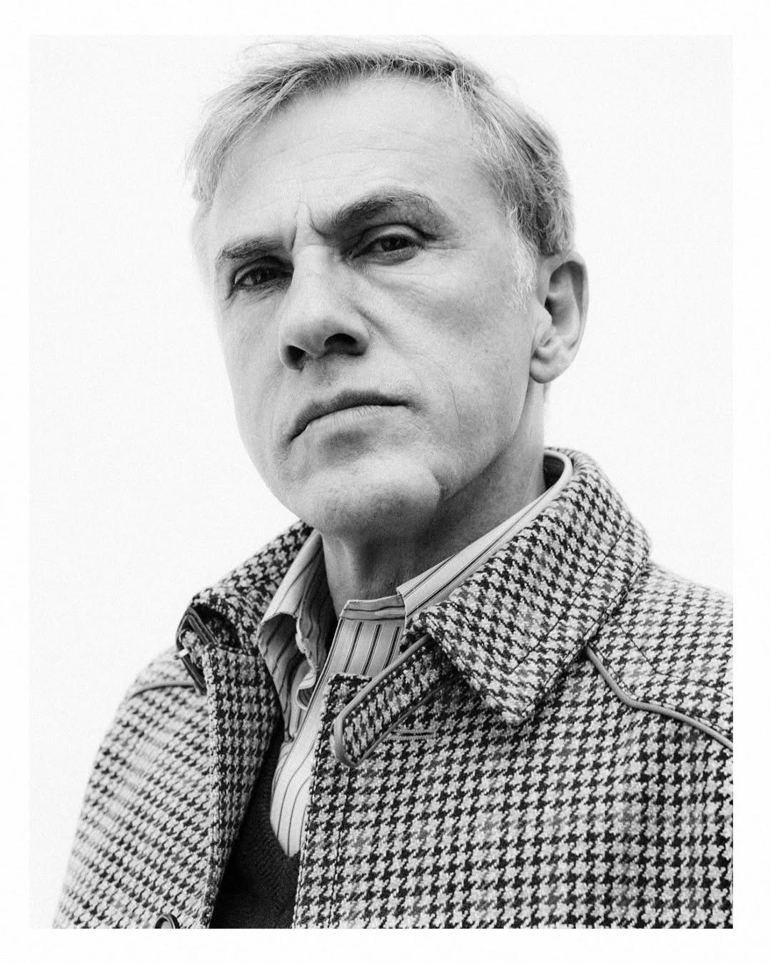 Black and white portrait of a mature man in a patterned coat, looking at the camera with a neutral expression, against a white backdrop.