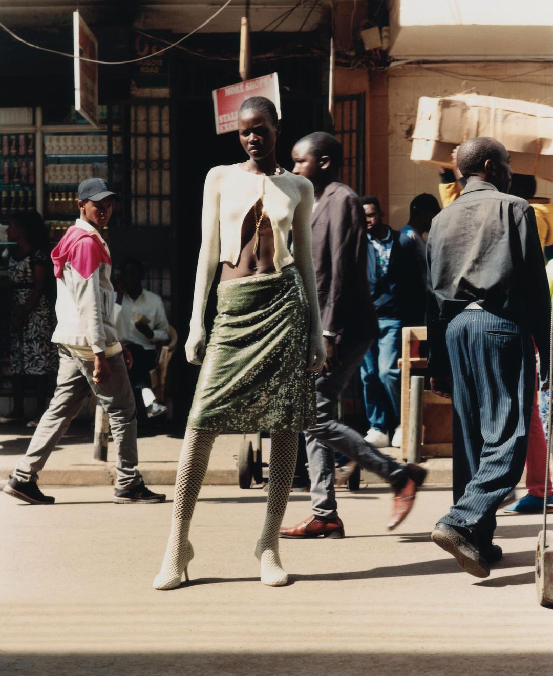 Fashion shot featuring a Black female model in a cream cardigan and green skirt, posing on a city street with people in the background.