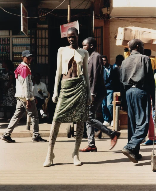 Fashion shot featuring a Black female model in a cream cardigan and green skirt, posing on a city street with people in the background.