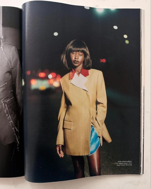 Fashion magazine photo of a woman in a tan blazer and blue shorts standing on a street at night with blurred lights in the background.