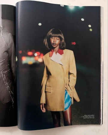 Fashion magazine photo of a woman in a tan blazer and blue shorts standing on a street at night with blurred lights in the background.