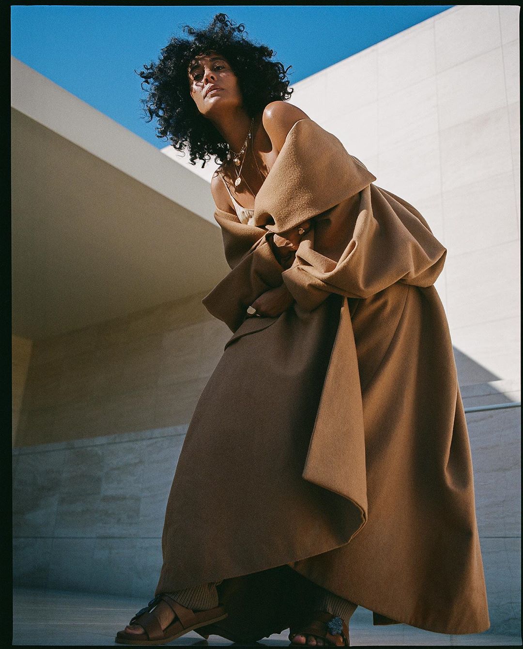 A woman with dark curly hair models in a brown coat and skirt in front of a neutral stone wall under a bright blue sky.