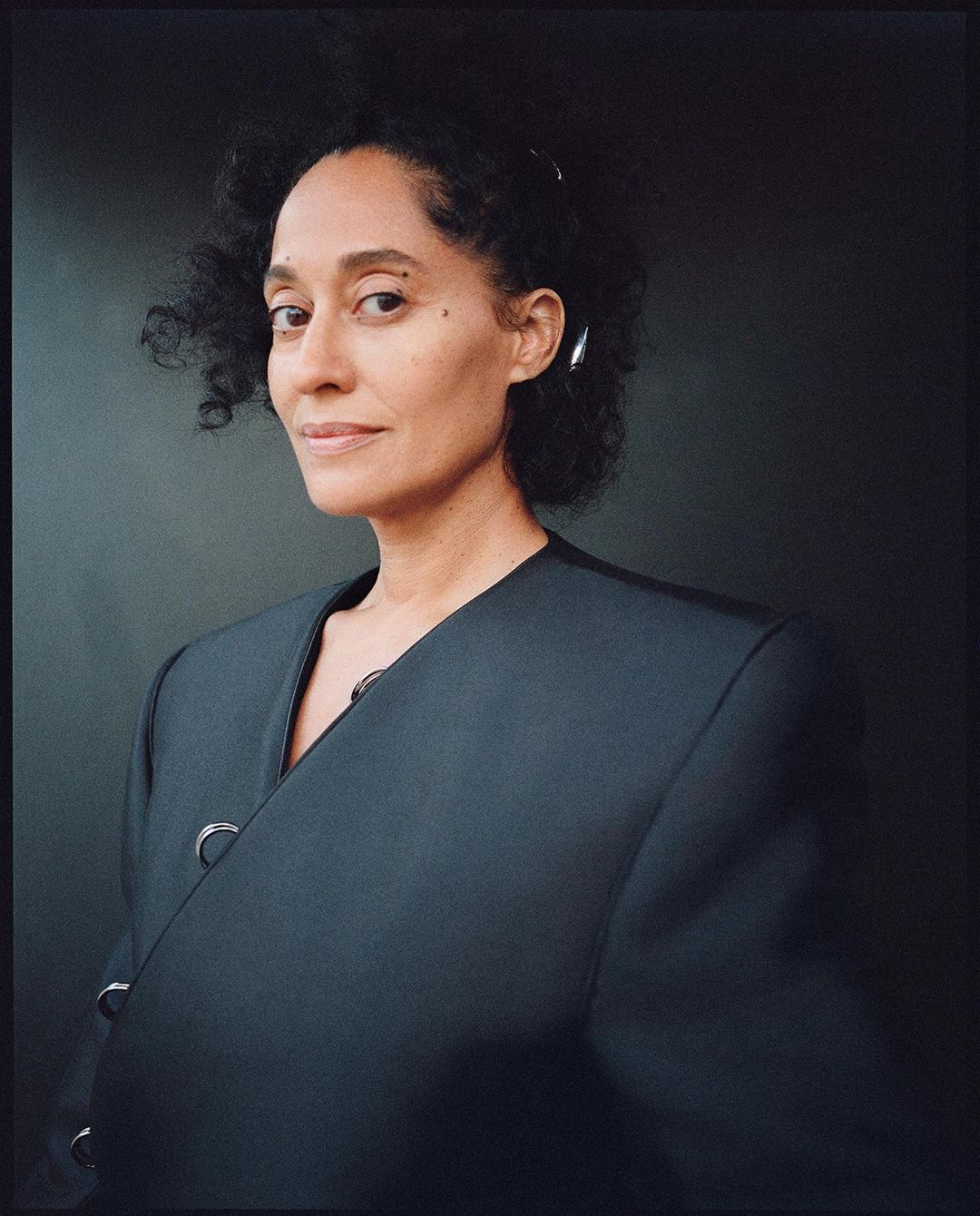 Portrait of adult woman with curly hair, wearing a black jacket, posing indoors in front of a dark gray background. Fashion shot.
