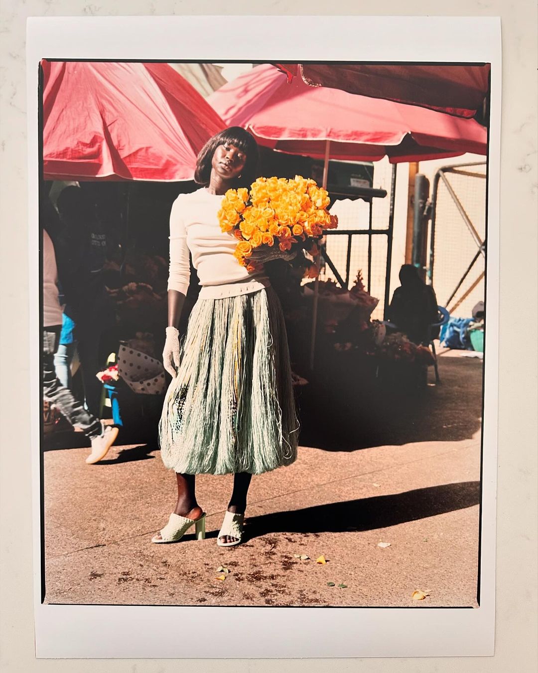 A striking image showcases a Black fashion model in an outdoor market, holding yellow roses and wearing a fringed skirt and gloves.