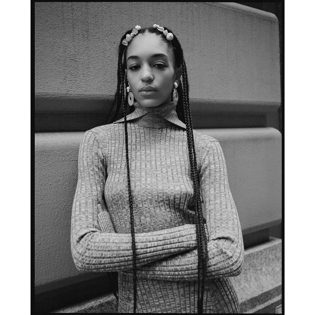 Monochrome portrait of an adult woman with braids and decorative hair beads, wearing a ribbed turtleneck and standing in front of a concrete wall.