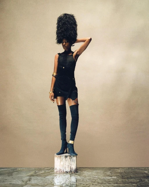 A woman with dark skin stands on a wooden block wearing a tall feathered hat, short black dress, thigh-high boots, and gold bracelets.