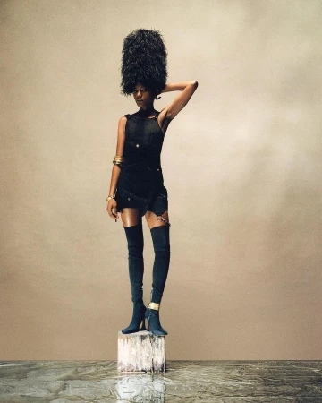 A woman with dark skin stands on a wooden block wearing a tall feathered hat, short black dress, thigh-high boots, and gold bracelets.