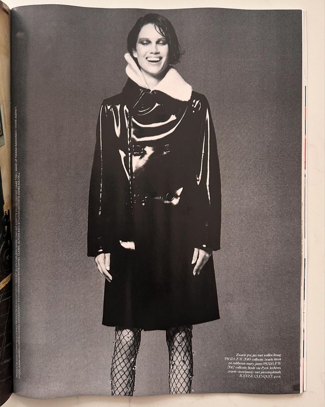 A smiling adult woman poses in a black, shiny coat with textured tights against a neutral grey background in a studio fashion shot.
