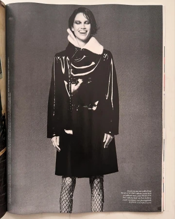 A smiling adult woman poses in a black, shiny coat with textured tights against a neutral grey background in a studio fashion shot.