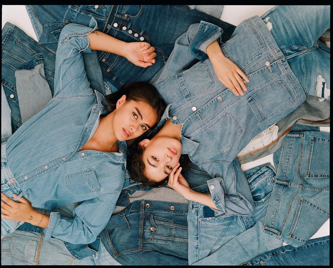 Two women lie on a pile of denim clothing, wearing denim jackets and shirts, illuminated by soft, natural light in a studio setting.
