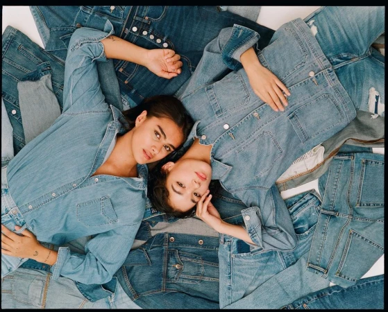 Two women lie on a pile of denim clothing, wearing denim jackets and shirts, illuminated by soft, natural light in a studio setting.