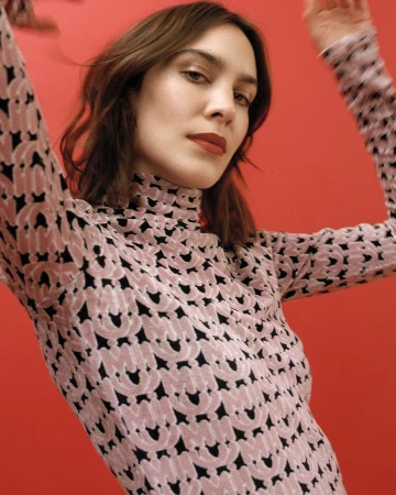 A woman wearing a pink patterned turtleneck top posing against a solid red background with her arms raised.