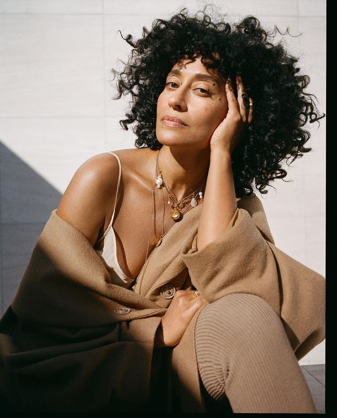 Portrait of a woman with curly hair wearing a tan outfit and jewelry, sitting and posing against a light wall in natural light.
