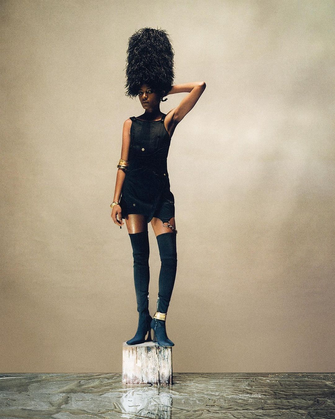 Fashion portrait of a woman in a black dress and ornate headpiece, standing on a podium in a studio setting with a neutral backdrop.