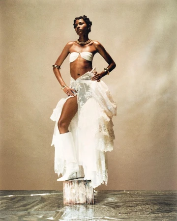 Fashion shot of an adult Black woman in a white outfit with jewelry, standing on a wooden post in shallow water against a neutral backdrop.