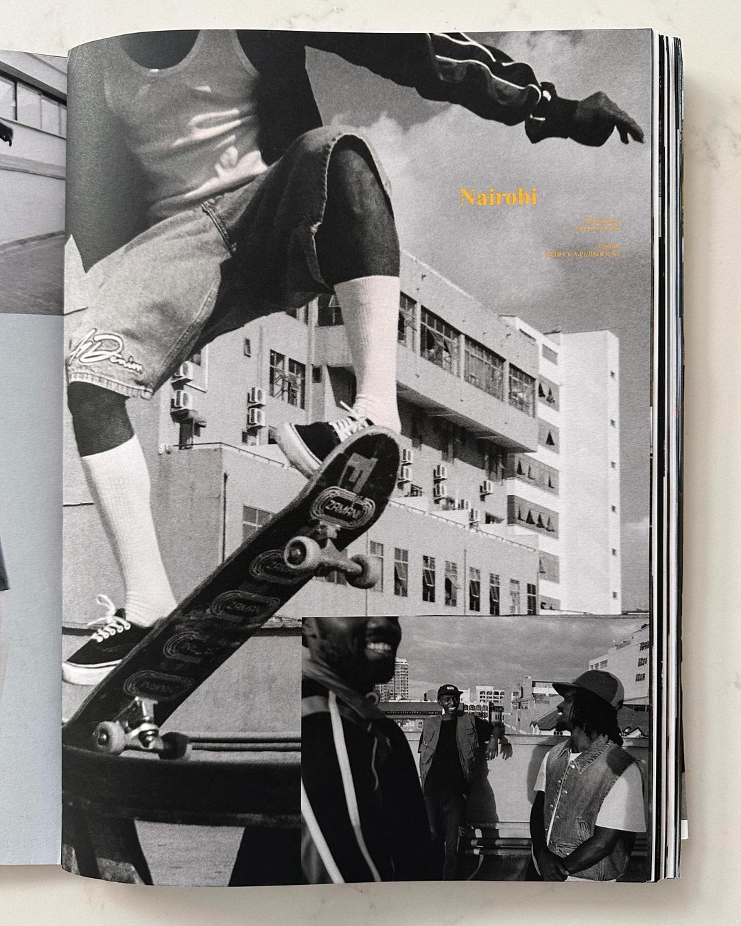 Black and white photo of a skateboarder performing a trick, with onlookers and urban architecture in the background, featured in a magazine.