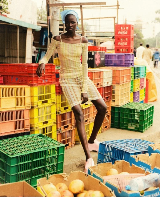 Woman in patterned dress poses in front of colorful crate stacks at an outdoor market, wearing a headscarf and light pink footwear.