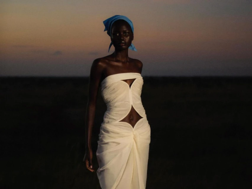 Woman in strapless dress and headscarf stands outdoors in a dusky field; fashion, beauty, and nature meet in a stylish portrait.
