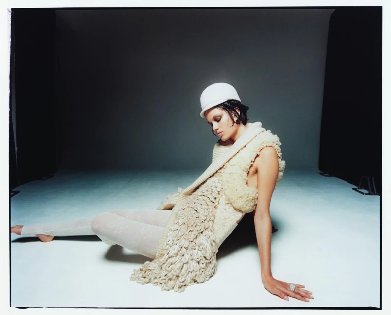A brunette woman wearing a white hat and textured shawl poses on a white floor in a photo studio with a dark background.