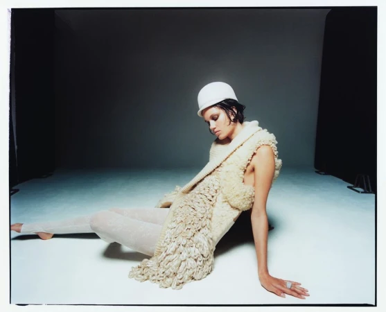 A brunette woman wearing a white hat and textured shawl poses on a white floor in a photo studio with a dark background.