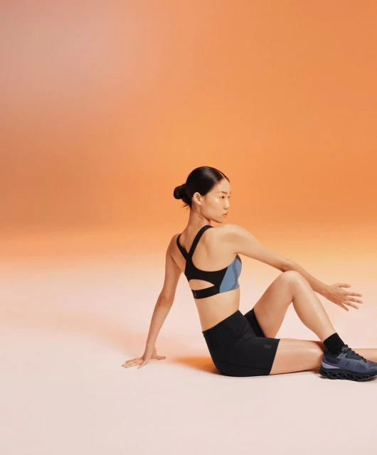 Asian woman in athletic wear sitting cross-legged in a twisting yoga pose against an orange gradient background in a fitness studio.