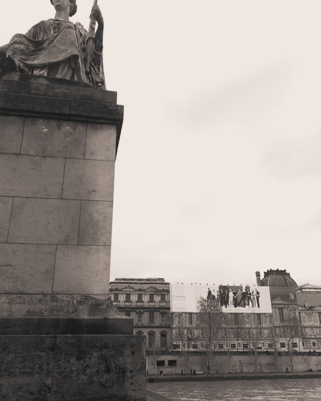 Monochrome architectural photograph featuring a classical stone statue on a pedestal overlooking a river and distant classical buildings under an overcast sky.