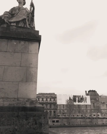 Monochrome architectural photograph featuring a classical stone statue on a pedestal overlooking a river and distant classical buildings under an overcast sky.