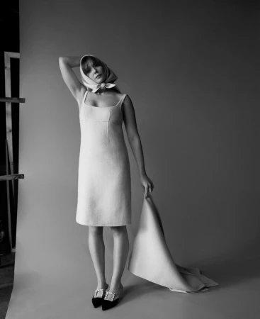 Monochrome portrait of a woman wearing a headscarf, a sleeveless dress, and jeweled mules against a grey backdrop.