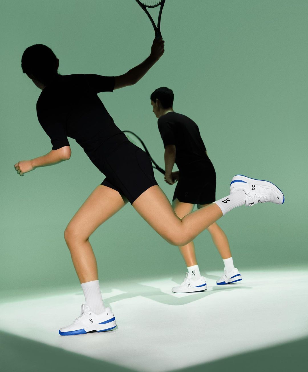 Two athletes run in white and blue tennis shoes with tennis rackets on a green background, showcasing speed and athletic apparel.