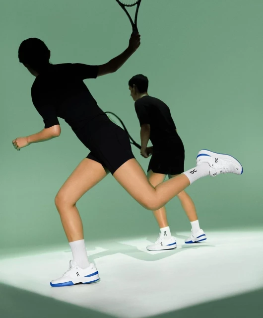 Two athletes run in white and blue tennis shoes with tennis rackets on a green background, showcasing speed and athletic apparel.