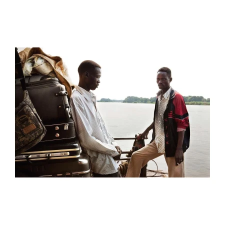 Two black men standing on a boat next to a stacked pile of luggage with a wide river in the background.