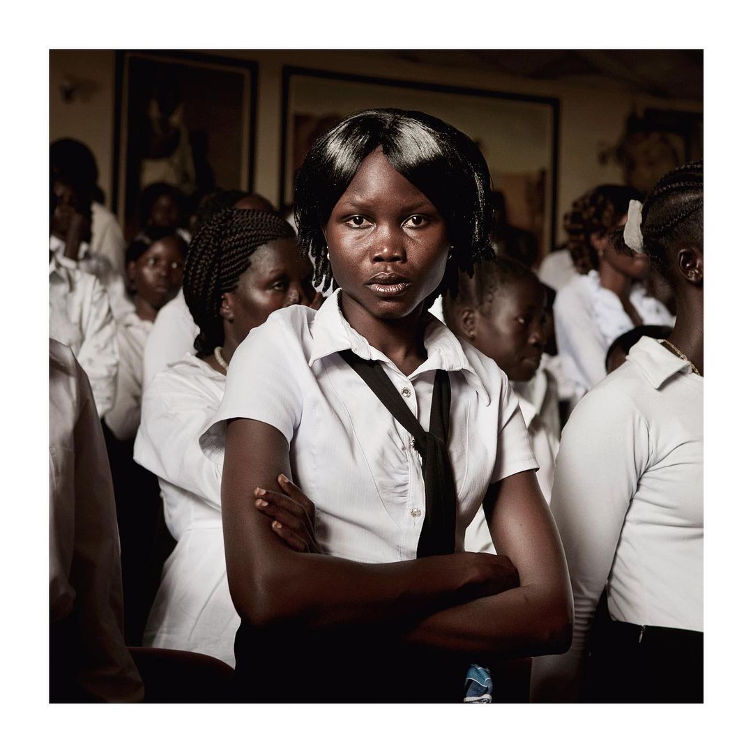 Portrait of a young Black woman in a school uniform, arms crossed, surrounded by a group of female students in a muted indoor setting.
