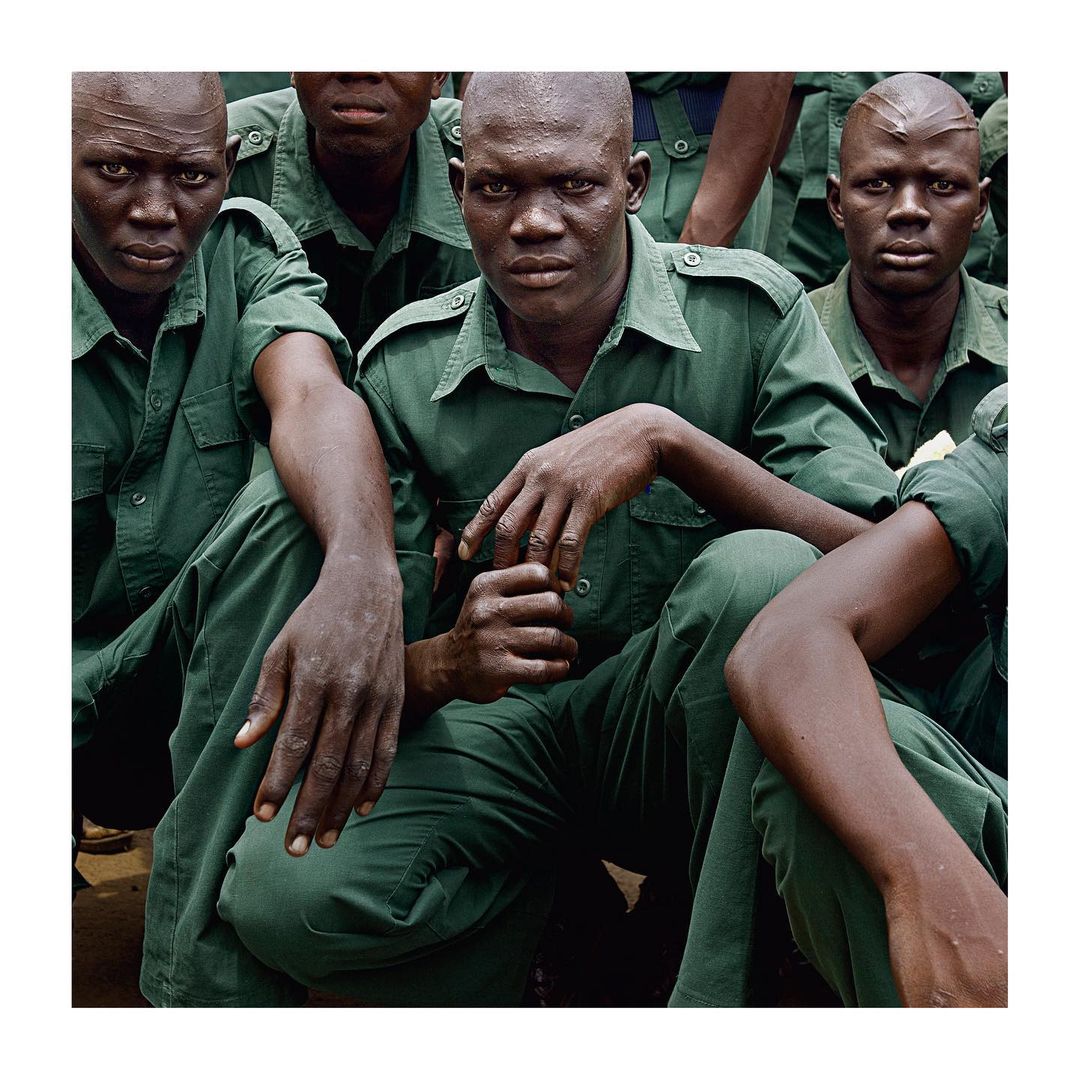 A group of African men in green uniforms, likely soldiers, are closely gathered, gazing directly forward with serious expressions in an outdoor setting.
